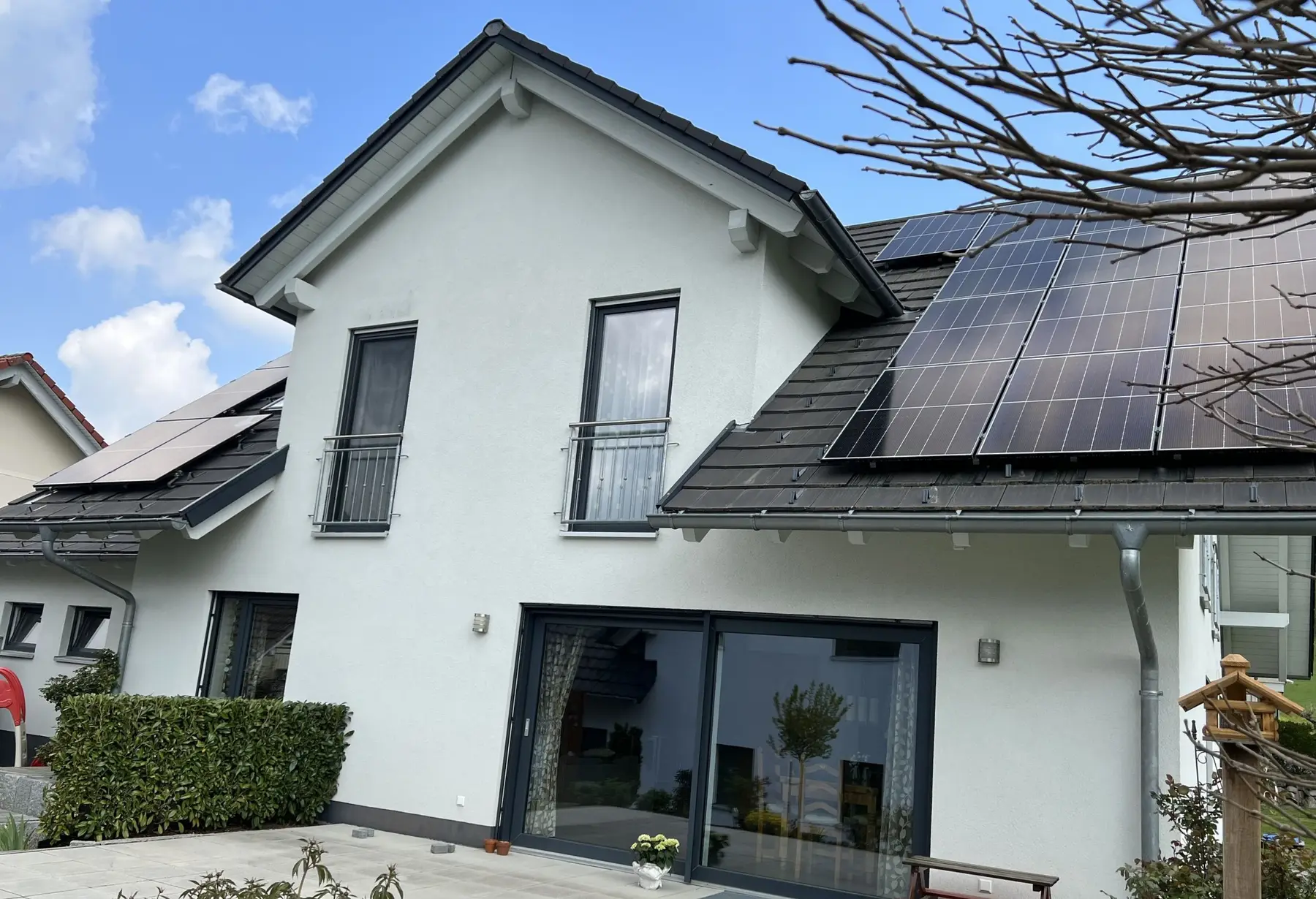 Weißes Einfamilienhaus mit dunklem Dach und schwarzen Solarmodulen, davor ein Vorgarten unter blauem Himmel