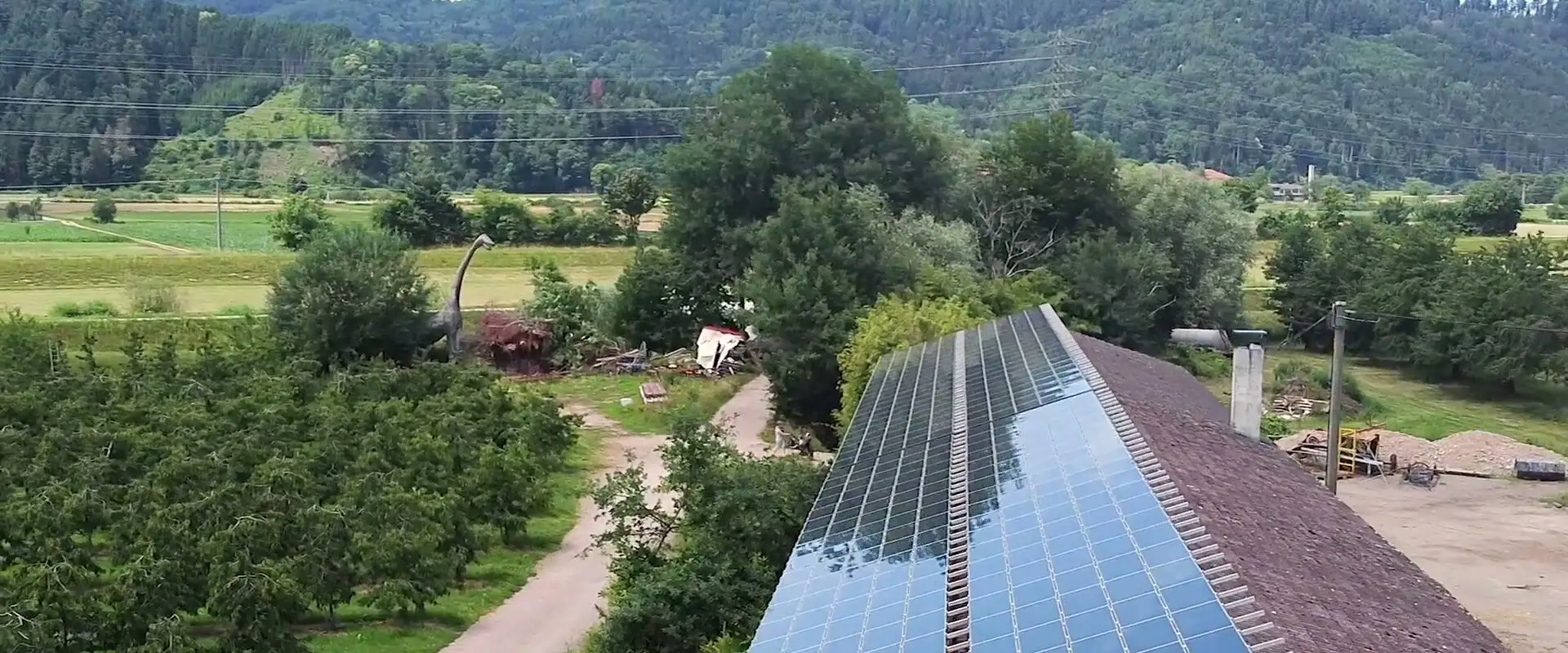 Luftaufnahme einer Scheune, deren Dachseite ganz mit Solarmodulen belegt ist, daneben Obstbäume, Felder und bewaldete Hänge