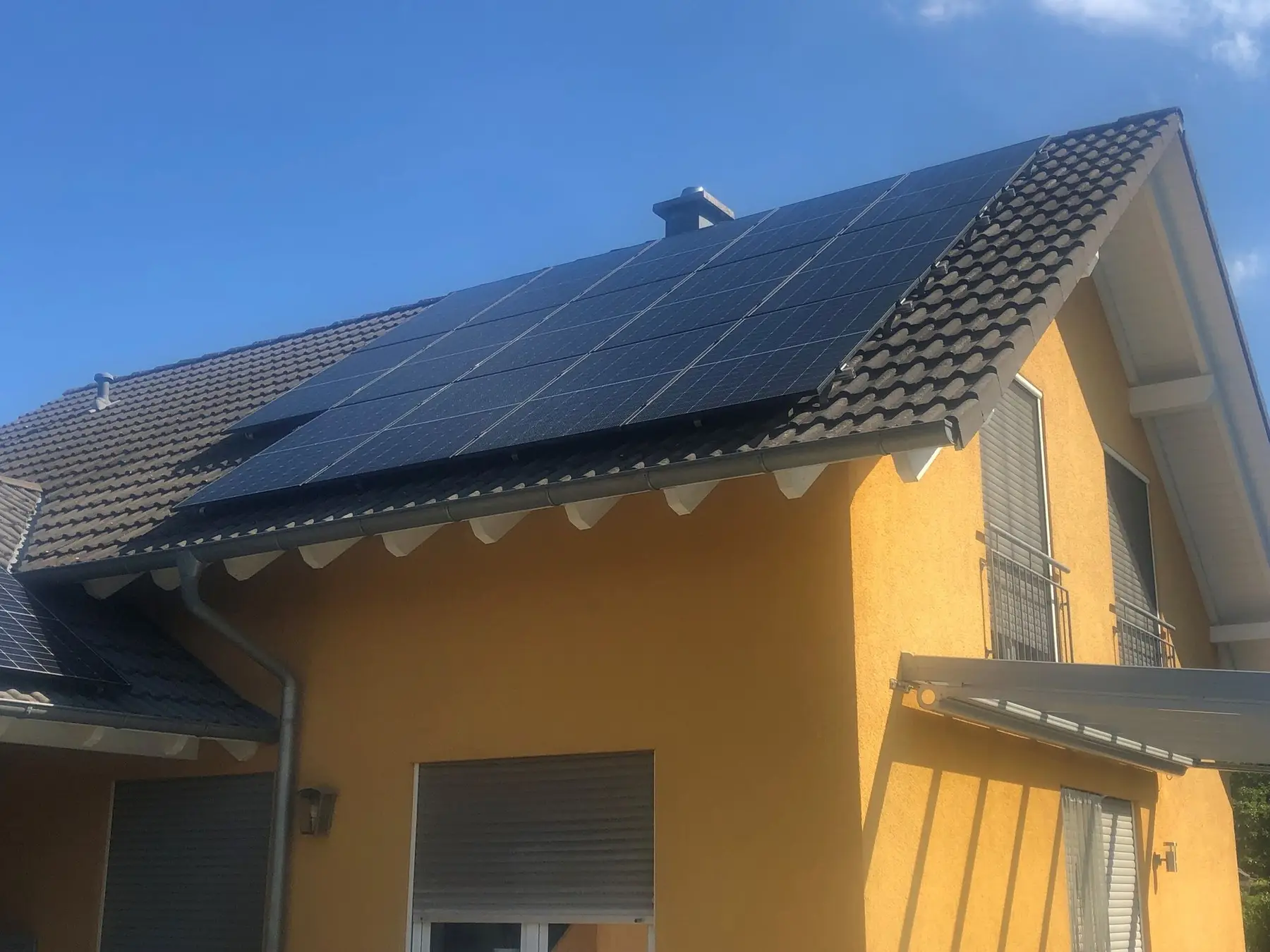 Einfamilienhaus mit gelber Fassade, auf dem braunen Ziegeldach schwarze Solarmodule vor blauem Himmel