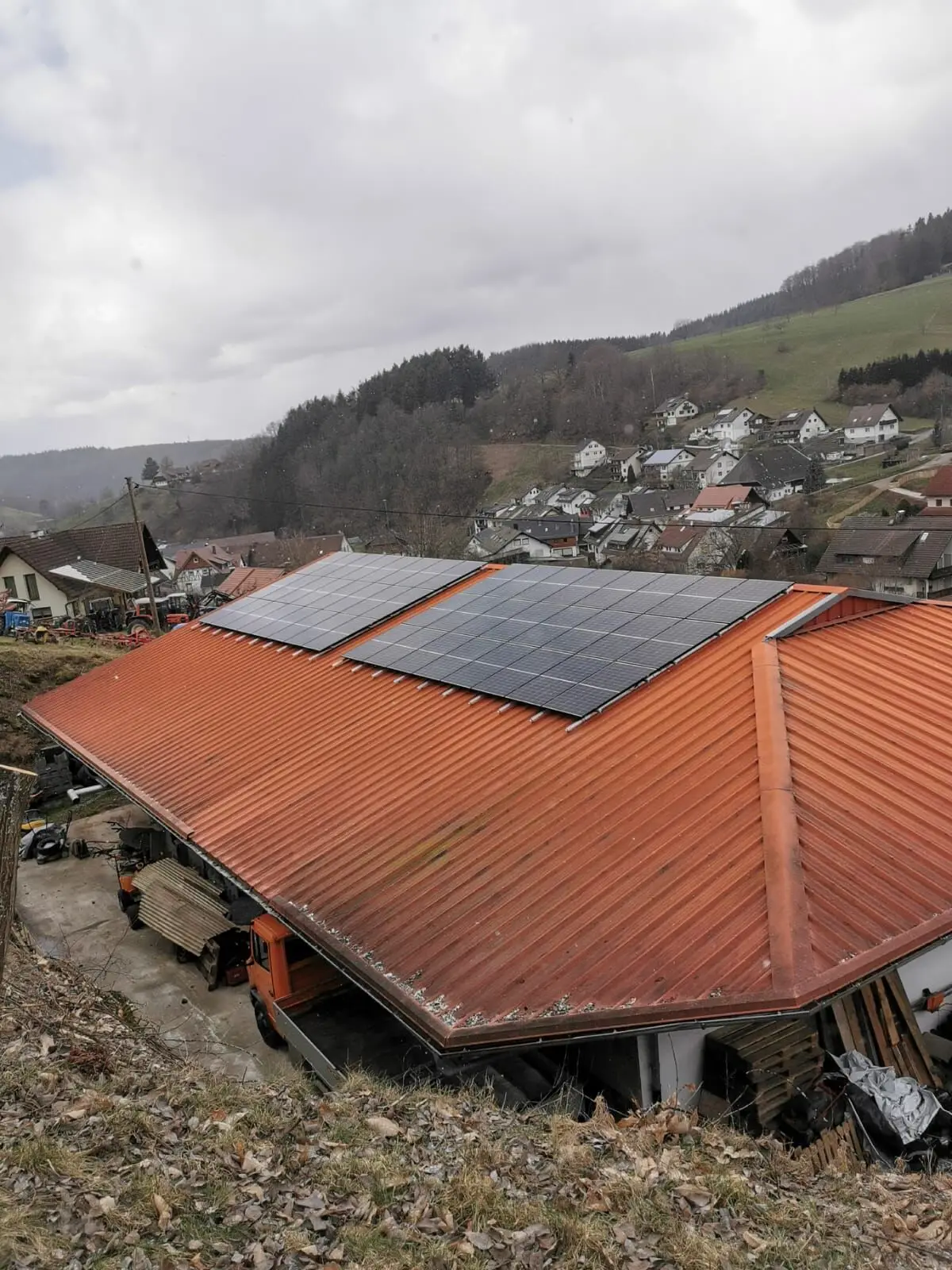 Zwei Modulfelder auf dem orangefarbenen Blechdach einer Halle, dahinter ein Dorf im Tal und bewaldete Hänge