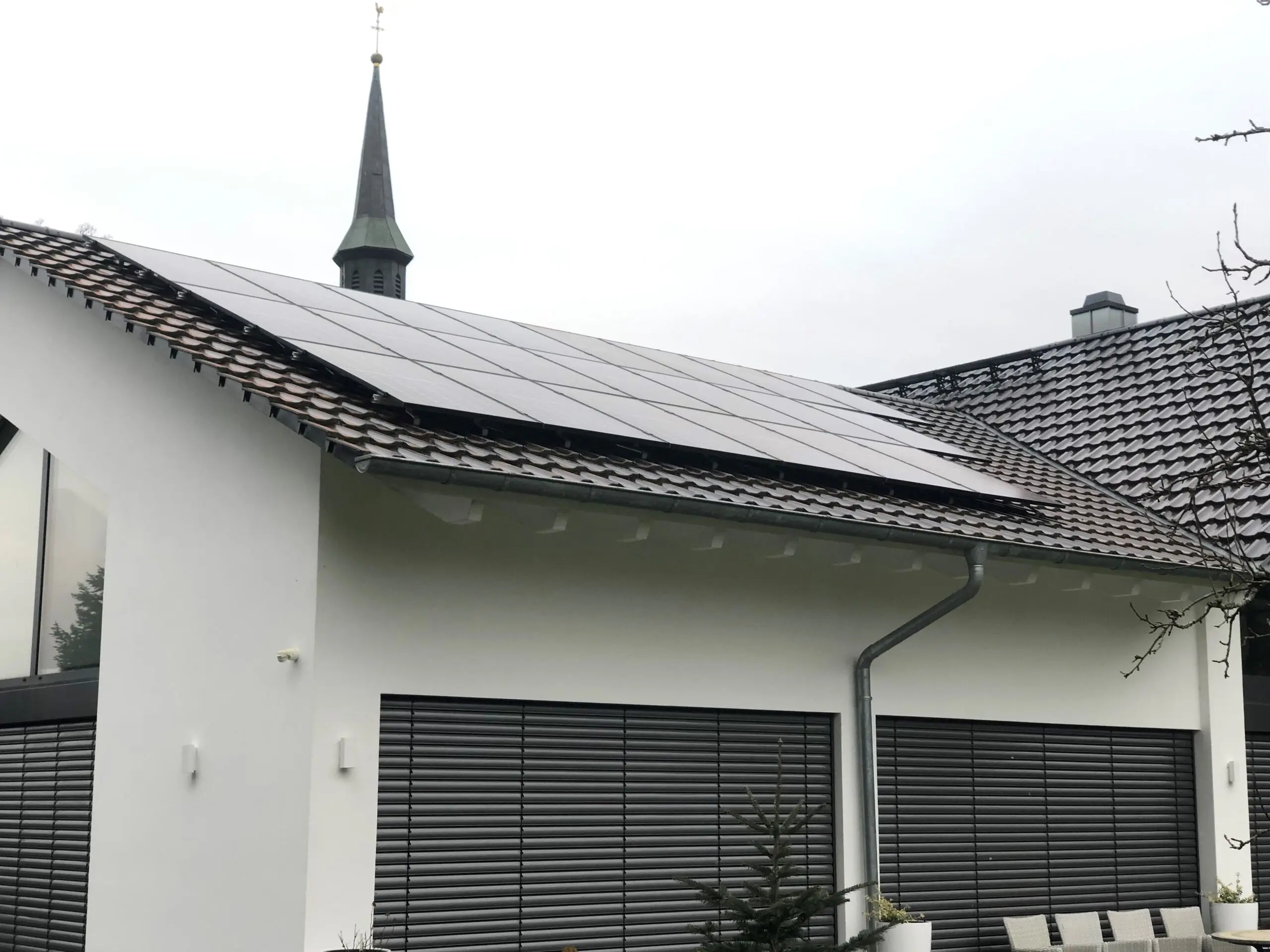 Wohnhaus mit weißer Fassade, auf dem Ziegeldach eine Photovoltaikanlage aus vollschwarzen Modulen, dahinter ein Kirchturm