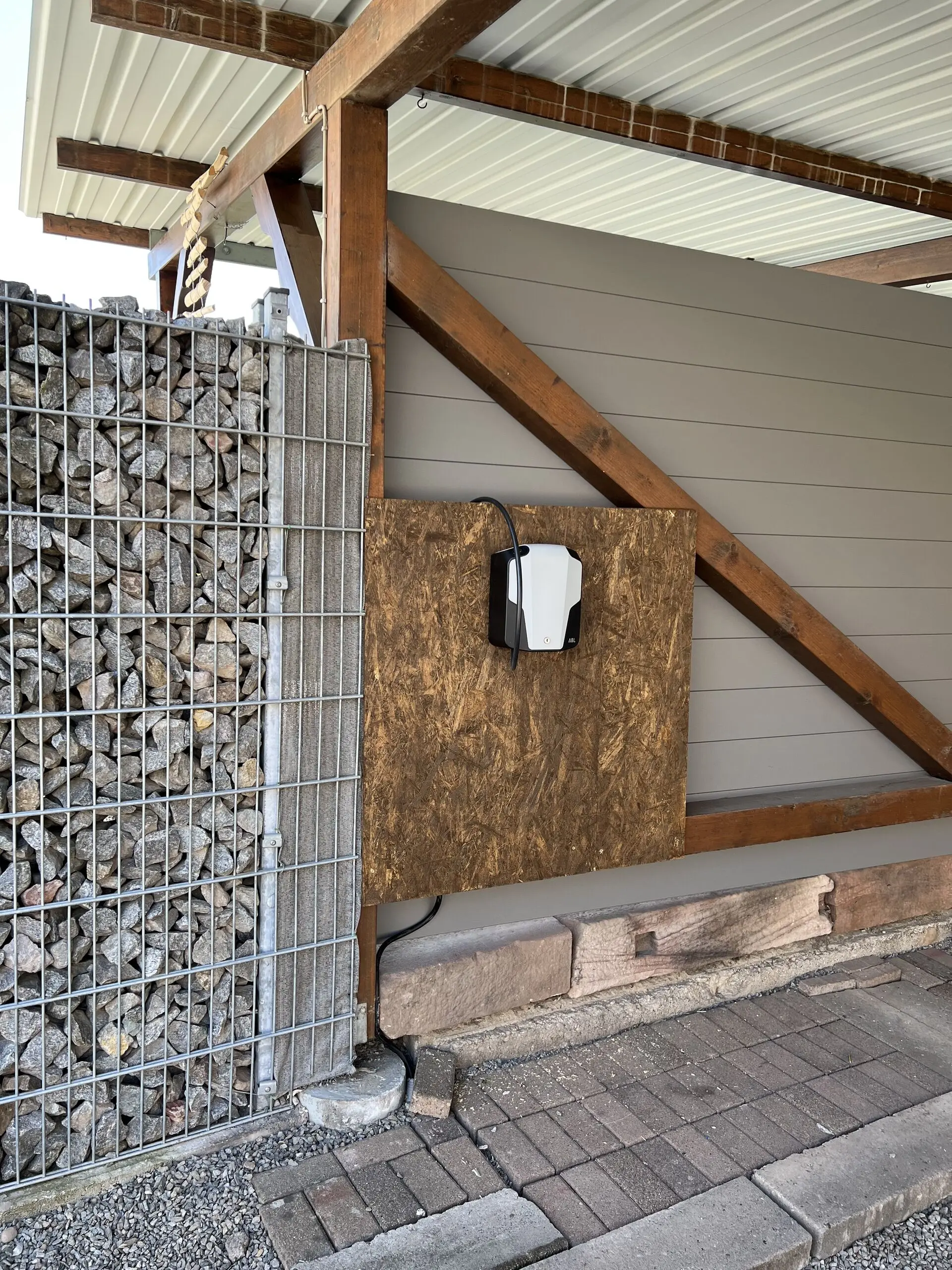 Weiße Wallbox an einer Holzplatte unter einem Carport, das Ladekabel führt an der Seite nach unten