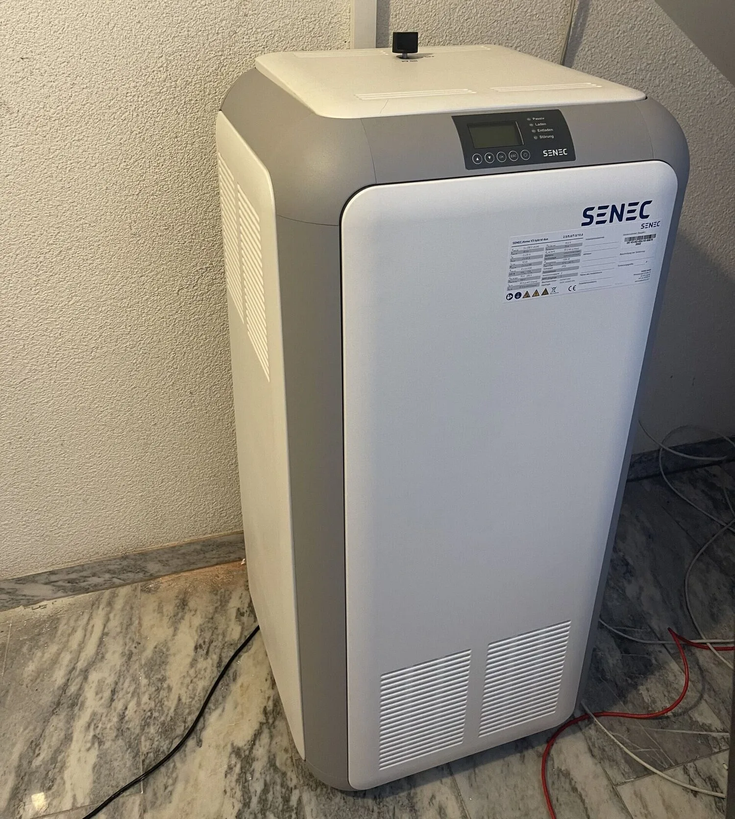SENEC-Stromspeicher in einem Hauswirtschaftsraum mit Display oben, Typenschild auf der Front und Anschlusskabeln am Boden